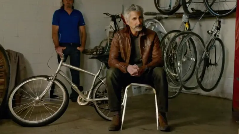 Un anciano encanecido con un jacket de cuero desgastado se sienta en una silla plegable delante de su bicicleta vieja en el centro de un garaje oscuro y sucio