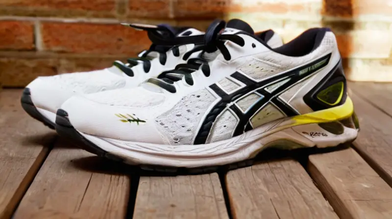 Un par de zapatos de correr negros y blancos con el logotipo ASICS descansan en un piso de madera junto a sus copias desgastadas, rodeados por un ambiente cálido e iluminado débilmente