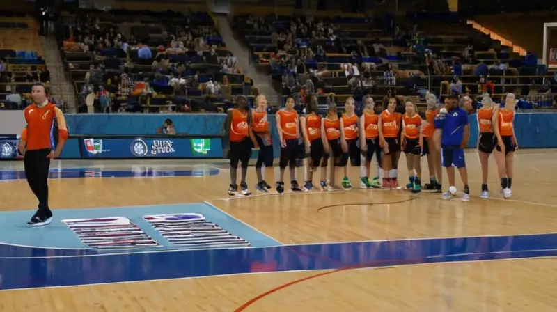 Un grupo de atletas (hombres y mujeres) vestidos con uniformes de pista cubierta se preparan para jugar baloncesto en una cancha con un tapizado de goma blanca y luces fluorescentes intensas