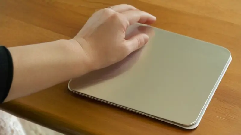 Una persona sostiene un dispositivo rectangular de plata con las manos apoyadas en la rodilla y los codos suaves sobre una mesa desvaída de madera