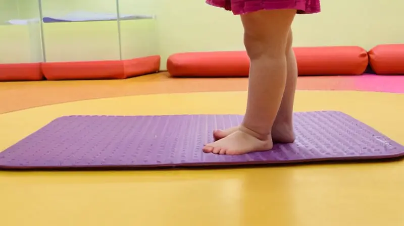 Una pequeña persona está sentada en el piso de una habitación de terapia iluminada donde hay almohadillas blandas de goma