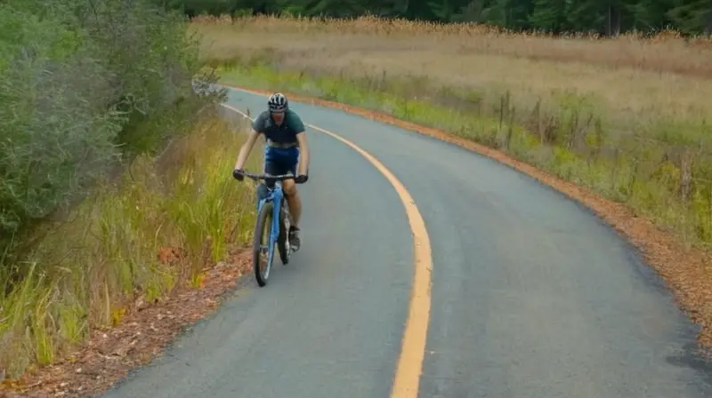 La escena muestra una carretera de asfalto desgastado y un camino de tierra con dos bicicletas apoyadas en un poste de madera