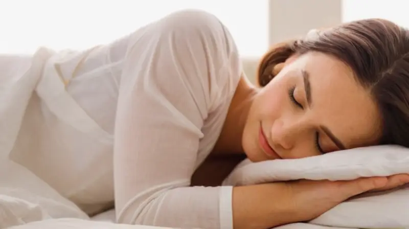 La mujer reposa en una cama con ropa blanca desgastada y sombreados calientes que sugieren un descanso sereno, aunque no exento de ansiedad